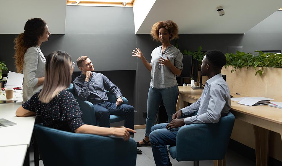 Five people are having a training meeting in a slightly dark office room. One colleague is stood up and is talking to the rest of her colleagues who are sat down and listening to her. 