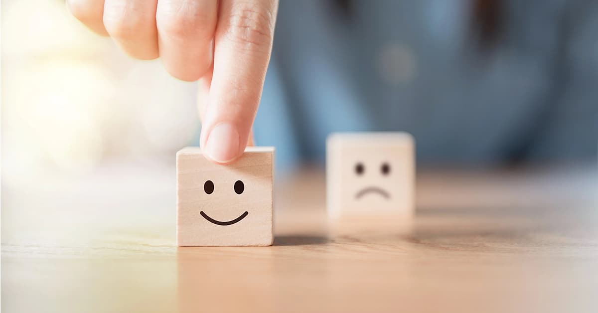 Two wooden blocks are on a table. One block is being held by finger and it has a happy face on. The other block is behind it and has a sad face.