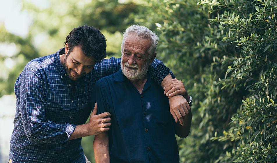 A man walks with his arm around his father alongside a green bush. They're smiling. 