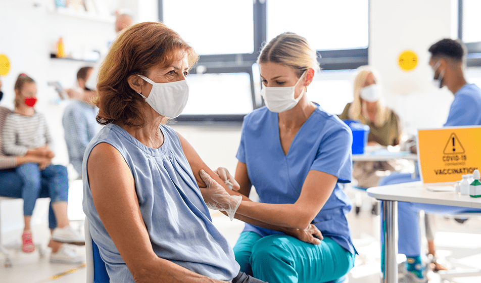 Two masked women sit, as one administers a vaccine into the other's left arm.