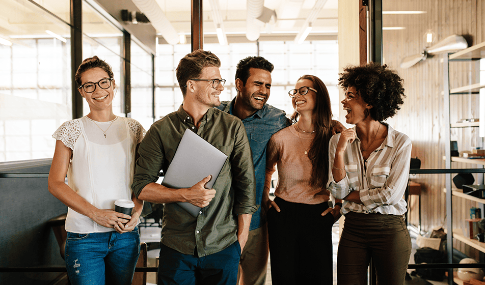 A group of five colleagues are standing together in their office and looking towards the camera. They are all laughing together.