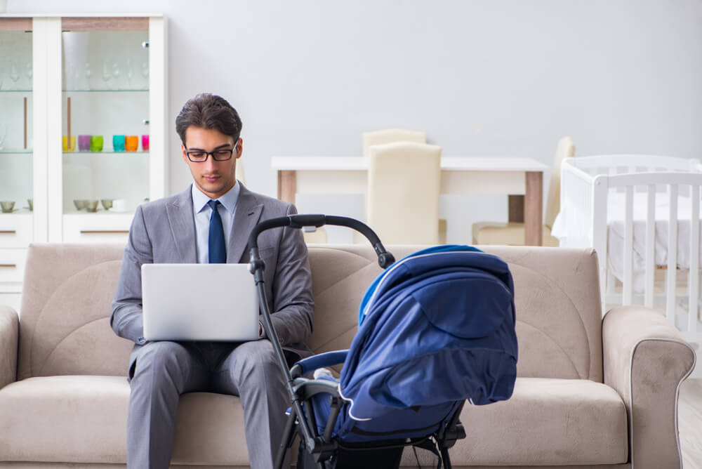A man in a suit is working from home and sat with his laptop on the sofa. Next to him is a blue pram as he is looking after his child whilst working.