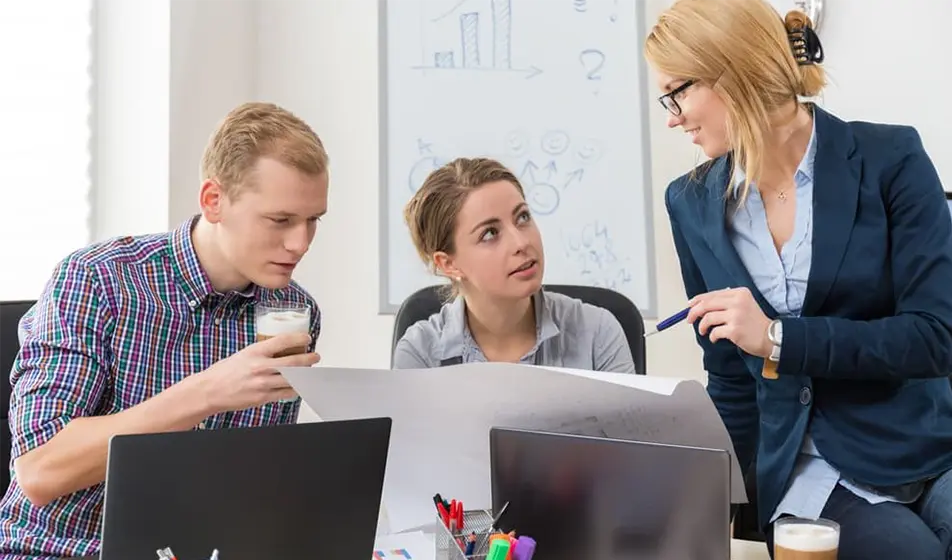 Three colleagues are sat at a desk together. They are have laptops in front of them but are all looking at a large piece of paper that has guidelines to project on.