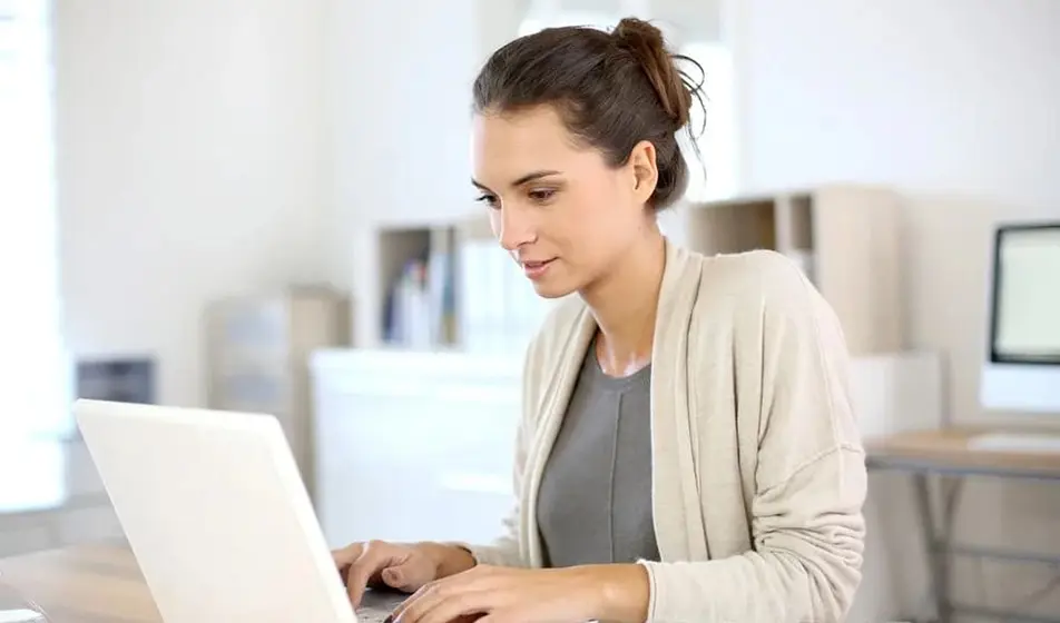 A woman with brown hair is wearing cream cardigan. She is working on her laptop and sat at a desk.