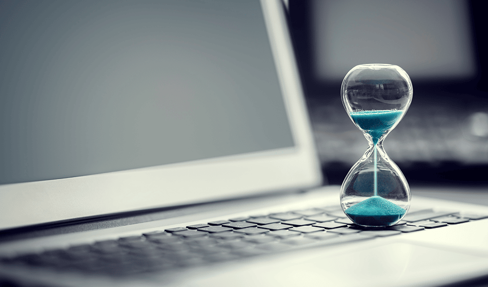 A sand timer with blue sand in it is standing on top of a laptop keyboard. It is about halfway through the timer.