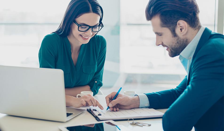 A man and a woman review a contract on a desk, with a laptop and ipad nearby. 